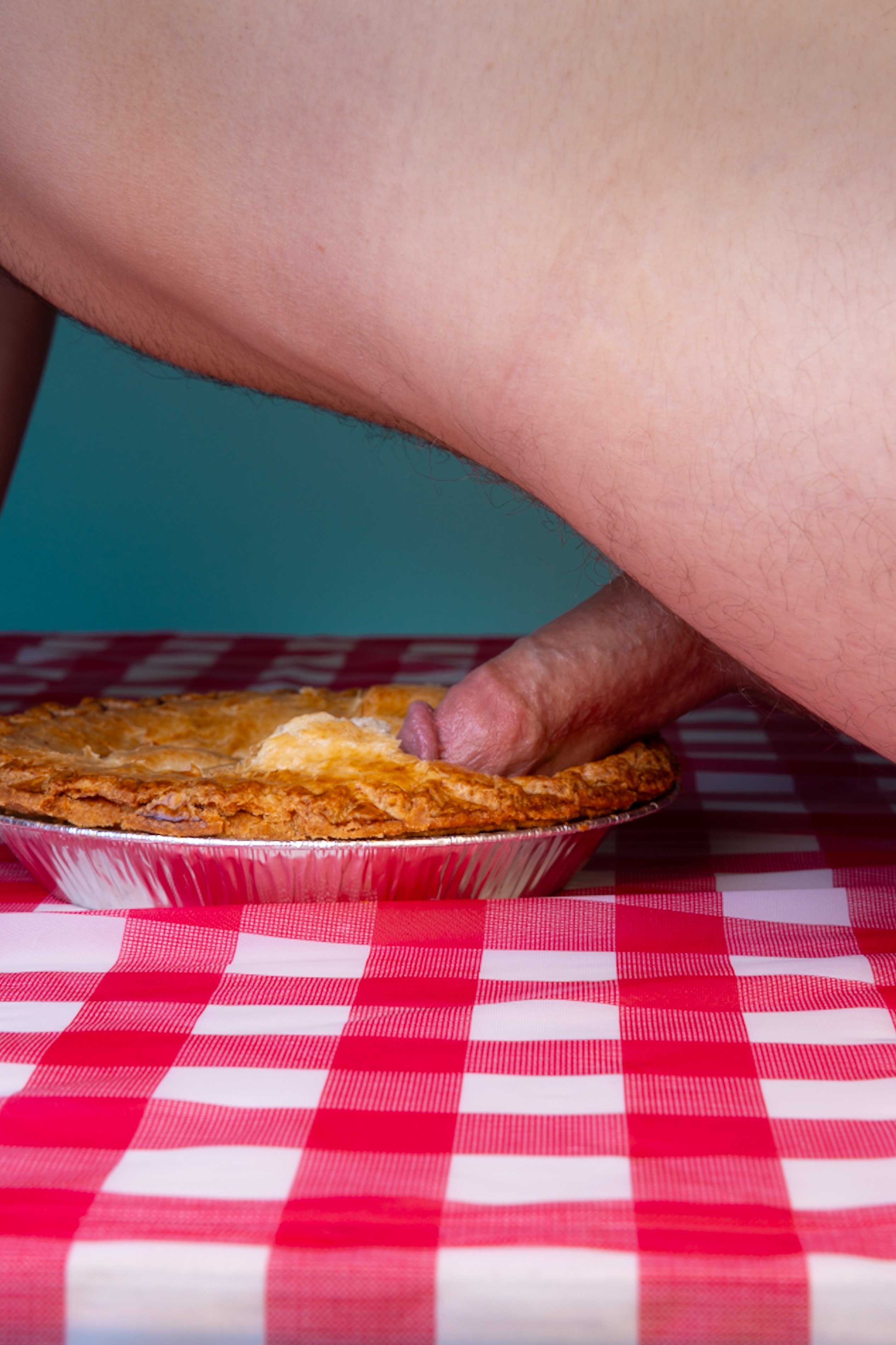 Erect penis pressed into apple pie on top of red checkered tablecloth, blue background. 
